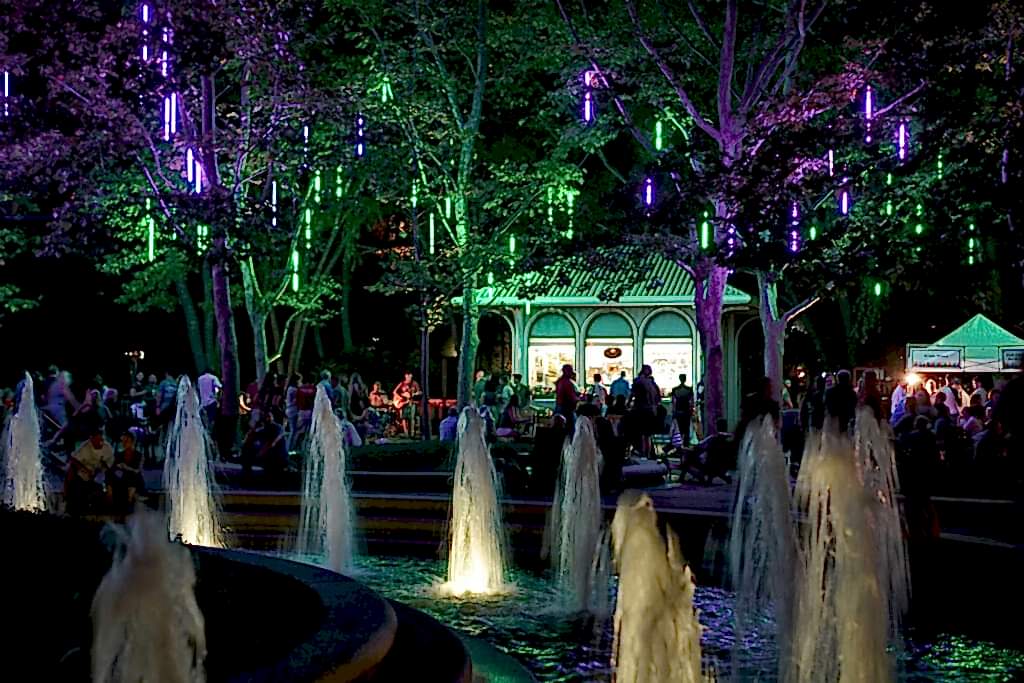 Trees lit purple and green over a plaza fountain at night